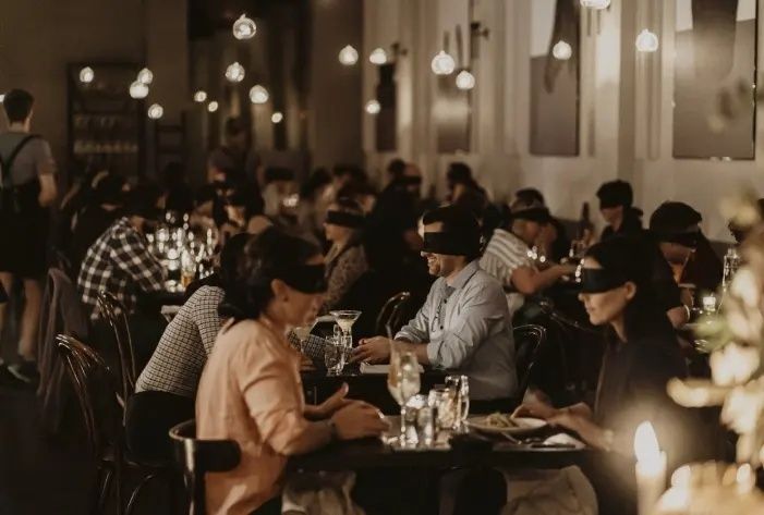 Gäste sitzen mit Augenbinden an gedeckten Tischen in einem Restaurant